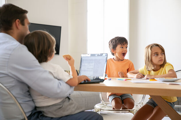 童年当爸爸在笔记本电脑上工作 抱着儿子跪在地上时 快乐的孩子们在画画退出孩子们在纸上画画高加索家庭坐在桌子旁童年 创造力和周末的概念女儿兄弟男人