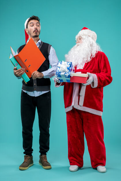 男人圣诞老人与年轻男性和礼物在蓝色墙上的正面视图套装正面音乐家