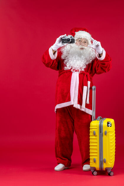 前面红墙上拿着相机的黄色袋子的圣诞老人的正视图节日圣诞圣诞老人