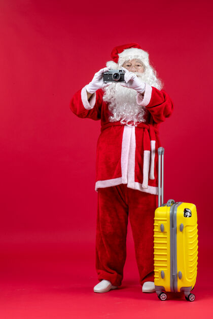 节日红墙上带黄包的圣诞老人用相机拍照的正面图拿着快乐包