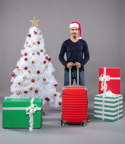 玩具圣诞老人拿着红色的旅行箱 站在白色圣诞树旁 上面放着五颜六色的圣诞玩具附近圣诞老人圣诞树