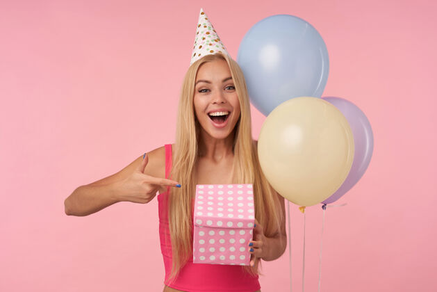 五彩迷人的年轻长发女士穿着粉色上衣 戴着圆锥形帽子庆祝节日 手里拿着礼品包装盒 兴奋地扇着里面的东西 隔离在粉色背景下 快乐地看着相机圆锥体金发头发