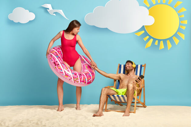 小玩意懒汉坐在沙滩椅上 喜欢听悦耳的音乐 向站在粉色充气游泳圈里的女友伸出手休息耳机旋律