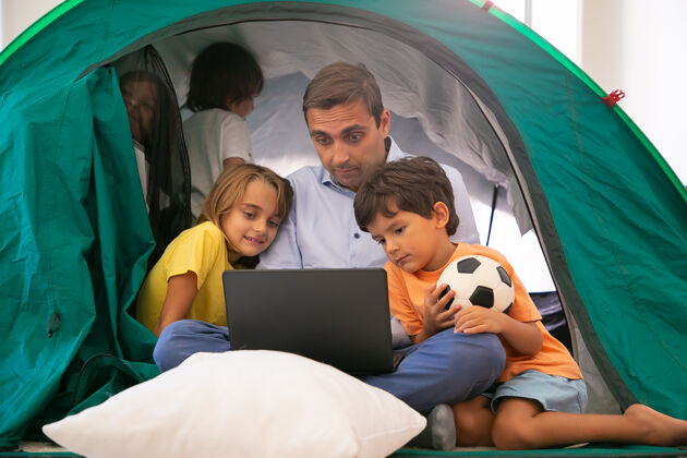 电影高加索爸爸盘腿坐在帐篷里和孩子们在家里 通过笔记本电脑看电影可爱的孩子拥抱爸爸 玩得开心童年 家庭时间和周末的概念孩子儿子父亲
