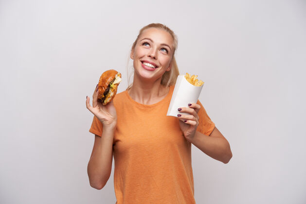 20多岁快乐迷人的年轻金发碧眼女性 手举汉堡包和炸薯条 开心地向上看 在白色背景下摆出大大的笑容长汉堡休闲
