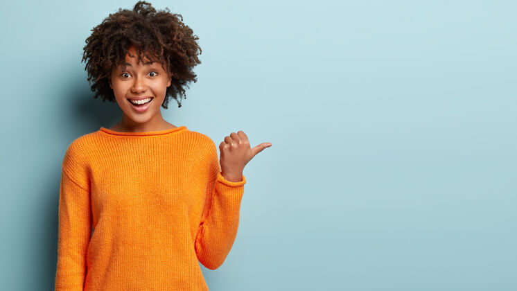 快乐照片中的一位快乐的美籍黑人妇女头发蓬松 指指点点 显示空白 高兴地为正在出售的商品做广告 身穿橙色套头衫 展示服装店的位置旁边卷发积极