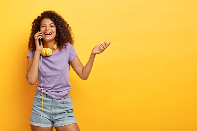 卷发积极的非洲裔美国女孩在手机上交谈的照片 笑容灿烂 举手 分享购物印象 讨论最后的时尚趋势交谈手机女性