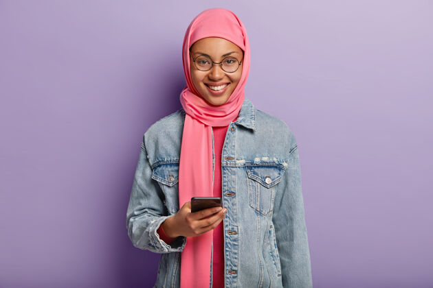 女士开朗的民族妇女 带着温柔的微笑 用手机在网上滚动新闻 读着有趣的邀请信 穿着粉色的hija 戴着圆眼镜 隔着紫色的墙与世隔绝民族头巾设备
