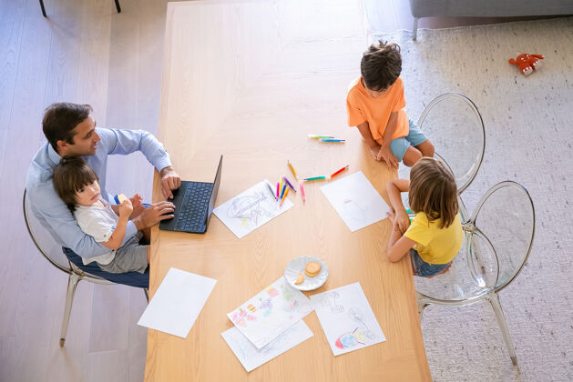 儿子俯视图：爸爸和孩子们一起坐在桌子旁弟弟妹妹用彩色钢笔涂鸦中年爸爸用笔记本电脑抱着小儿子童年 周末和家庭的概念可爱绘画孩子
