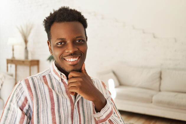 英俊快乐积极的年轻非洲裔美国男性坐在室内 手放在下巴上的肖像积极现代表情
