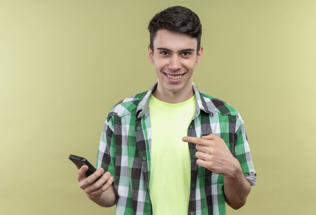 家伙一个穿着绿色衬衫的白人年轻人微笑着指着他手上的手机 背景是孤立的绿色微笑点绿色