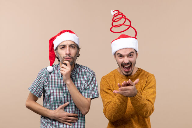 好奇的前视图两个家伙一个好奇的和一个快乐的圣诞老人帽子在米色孤立的背景两个帽子伙计们