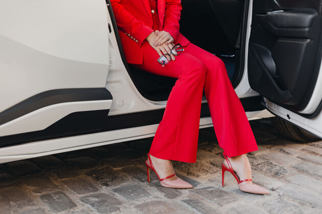 高穿着高跟鞋和手拿眼镜的性感富商美女穿着红色西装在白色汽车里摆姿势女士西装女人