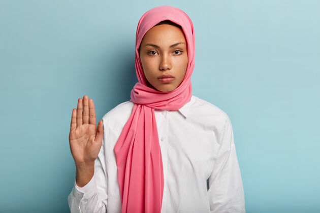 女人慢下来 停下来严肃的深色皮肤的阿拉伯女人表现出拒绝的姿态 向她举起手掌 表示拒绝 戴着粉色的头饰和白色的衬衫 隔离在蓝色的墙上头巾感觉干扰