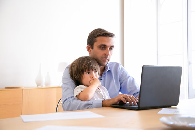 可爱严肃的父亲用笔记本电脑上网 抱着可爱的儿子白人中年男子穿着衬衫 和孩子坐在桌子旁 看着屏幕 工作父亲和家庭的概念看笔记本电脑屏幕