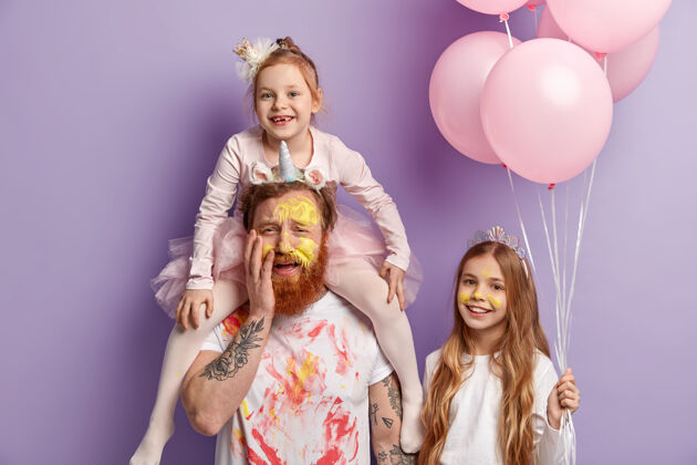 父亲三个家庭成员在紫色的墙壁上摆出室内姿势有趣的两个女儿和爸爸玩得很开心 用五颜六色的水彩画给脸上色 一起庆祝儿童节娱乐理念满足绘画狐狸