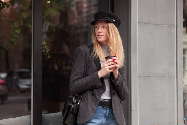 休闲体贴迷人的年轻女子 金色长发 一边喝着咖啡 一边走在户外 一边沉思着 身穿黑色外套 戴着黑色帽子头饰情感表情