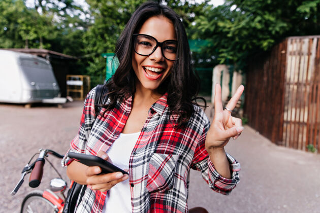 街道快乐的戴眼镜的白人女孩在城市里表达积极的情绪穿着格子衬衫手持智能手机的迷人女人的户外照片欧洲活跃时髦