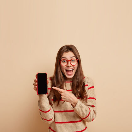 站立快乐的高加索人女性的垂直镜头指向智能手机设备 为你的广告文字显示黑色的空白屏幕 笑 有愉快的表情 隔离在米色的墙壁和自由的空间眼镜在线设备