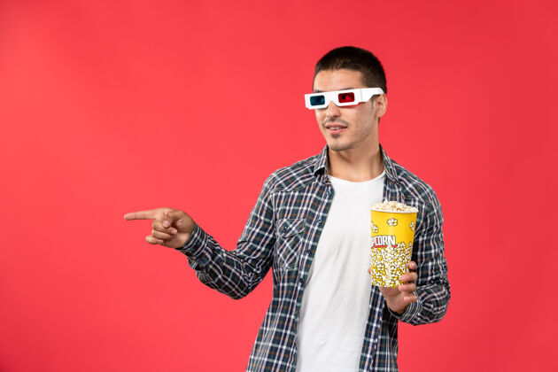 观点正面图戴着d型太阳镜的年轻男性拿着爆米花包在浅红色的墙上看男性电影电影院男性爆米花