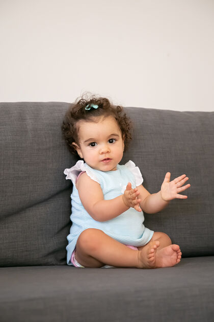 室内可爱的深色卷发女婴 穿着淡蓝色的布 坐在家里的灰色沙发上 一副和蔼可亲 拍手叫好的样子家里的孩子和童年的概念幼儿鼓掌可爱