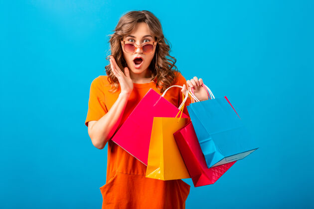 包图片中的兴奋迷人的微笑时尚女性购物狂穿着橙色的新潮连衣裙拿着购物袋在蓝色的工作室背景隔离购买蓝色持有