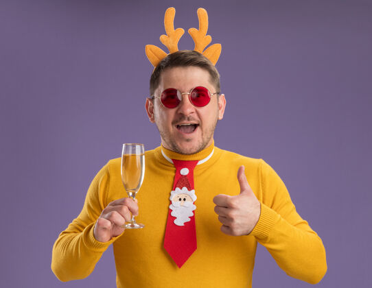 圣诞节穿着黄色高领毛衣 戴着红色眼镜的年轻人 戴着有趣的红色领带 戴着鹿角边 手里拿着一杯香槟 高兴而愉快地微笑着 竖起大拇指站着微笑站着穿