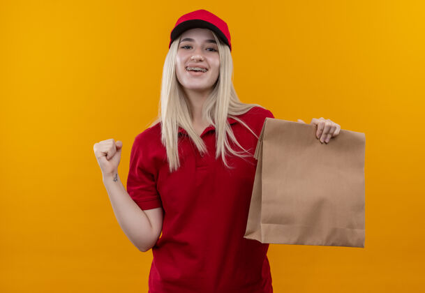 支撑微笑的小女孩穿着红色t恤 戴着帽子 戴着牙套 拿着纸袋 在孤立的橙色背景上显示“是”的手势穿着拿着送货