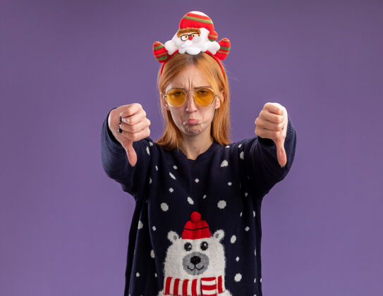 眼镜悲伤的年轻漂亮女孩穿着圣诞毛衣 戴着圣诞发箍 戴着眼镜 拇指朝下 在紫色背景上与世隔绝显示悲伤毛衣