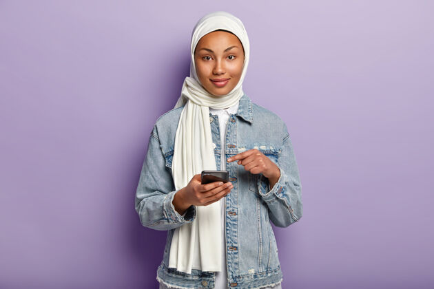 手机黑皮肤的快乐混血女孤立镜头 追随宗教 指着手机屏幕 要求在网站上看网络新闻 孤立在紫色的墙上头巾短信手机