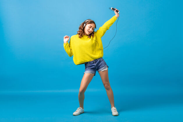 运动迷人的笑容可掬的快乐女人 戴着耳机 穿着时髦的服装 在蓝色的工作室背景下 穿着短裤和黄色的毛衣 一边跳舞一边听音乐微笑女人明亮