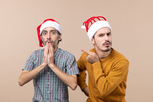 好奇正面图两个好奇的家伙戴着圣诞帽 一只手放在下巴上 另一只手指在米色孤立的背景上展示他的朋友男性显示手