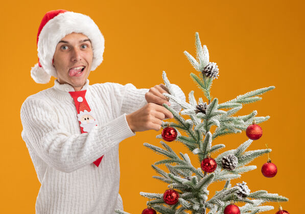舌头可爱的年轻帅哥戴着圣诞帽打着圣诞领带站在圣诞树旁用圣诞球装饰圣诞树领带顽皮快乐