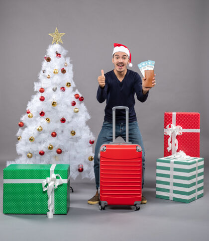 圣诞老人一个拿着红色手提箱的人高兴地展示了他的旅行票 并在灰色上做了一个竖起大拇指的标志圣诞帽礼物圣诞树