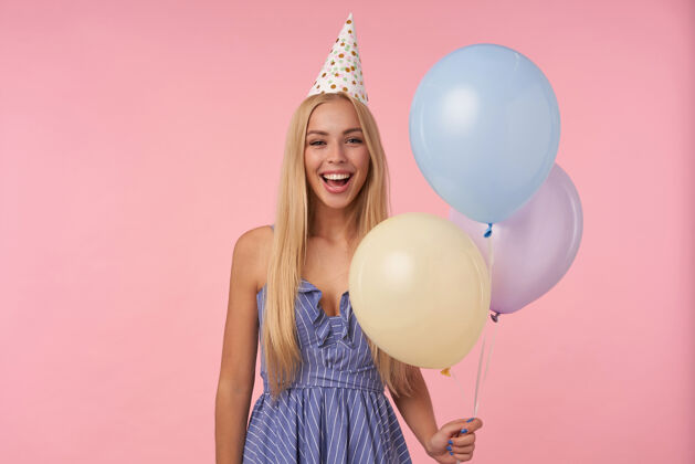 帽子快乐的年轻长发女性在五颜六色的气球里摆姿势 穿着蓝色的夏装和生日帽 庆祝节日 隔离在粉色的背景下长发头发迷人