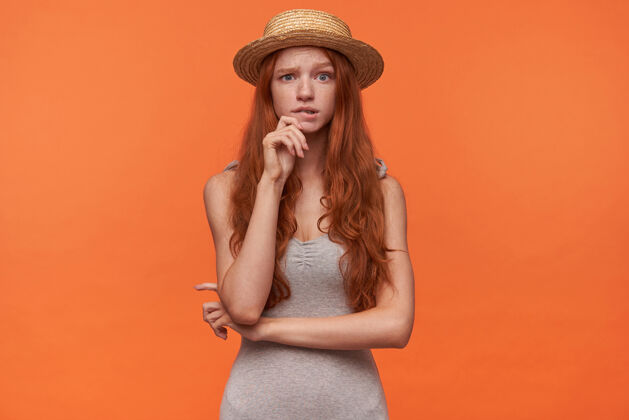 怀疑困惑的美丽年轻女性 卷曲的狐狸尾巴 穿着休闲服 戴着灰色衬衫草帽 站在橙色背景上 手放在下巴上 疑惑地看着镜头困惑自然衬衫