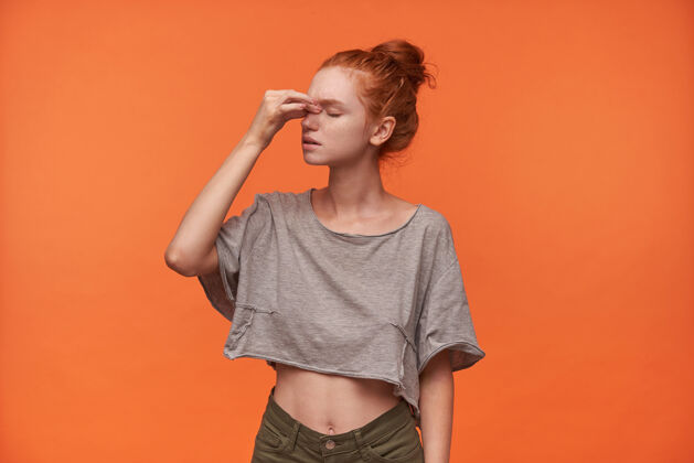 眼睛摄影棚照片中 一位穿着休闲服的年轻女子手拿鼻梁 闭着眼睛在橙色背景上摆姿势 红发打结手结面部