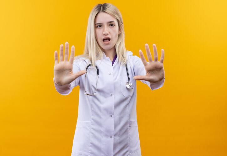 停止悲伤的医生年轻女孩穿着医用长袍戴着听诊器 双手在孤立的黄色背景上做着停止的手势穿着长袍医疗