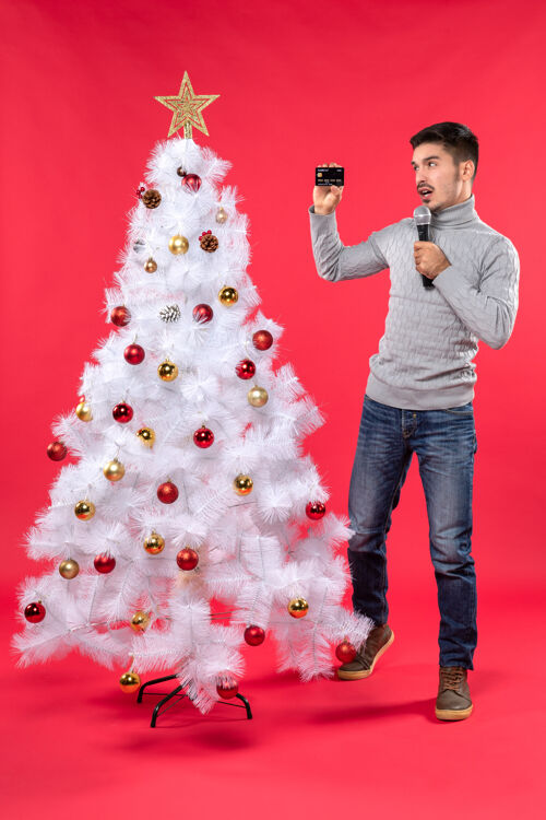 自信的家伙圣诞气氛 自信的家伙穿着牛仔裤站在装饰好的圣诞树旁 拿着麦克风给大家拍照拿着圣诞微笑