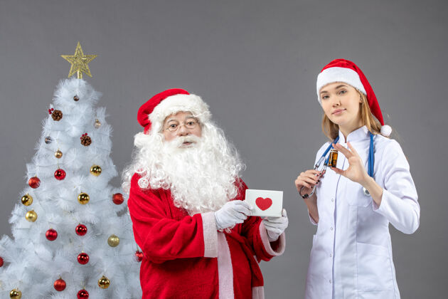 十二月圣诞老人和女医生的前视图 女医生拿着装着药片的小瓶子在灰色的墙上快乐女性快乐