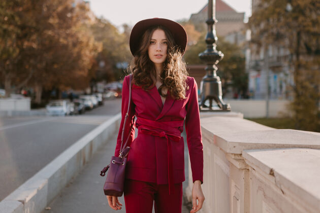 户外穿着紫色西装的漂亮时髦女人走在城市街头 春夏秋冬时节时尚潮流戴着帽子 手拿钱包秋天女士配饰