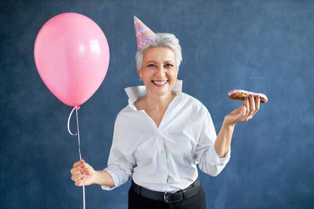 老年无忧无虑的快乐中年妇女 穿着优雅的衣服 戴着粉红色的圆锥形帽子 手里拿着空气和氦气球 面带微笑 在聚会上玩得很开心快乐60岁开朗