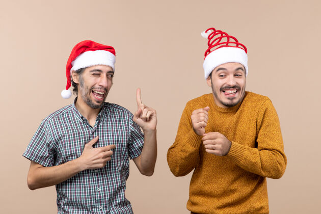伙计们正面图两个戴着圣诞帽的快乐的家伙一个眨着眼睛在米色孤立的背景上两个帽子微笑