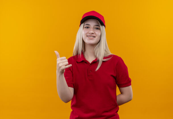 电话微笑送货的年轻女孩穿着红色t恤 戴着戴着牙套的帽子 在孤立的橙色背景上显示电话手势红色支架女孩