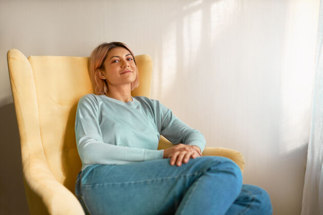 室内可爱无忧无虑的年轻白人女性穿着休闲服坐在白色窗帘上的黄色扶手椅上现代休息平静