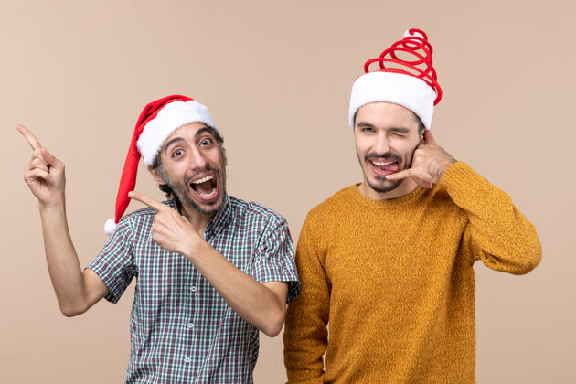 前面前视图两个带着圣诞帽的微笑的家伙 一个在展示什么 另一个在米色孤立的背景上打电话给我视图圣诞老人两个微笑的家伙