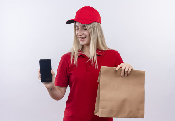 微笑微笑送货的年轻女孩穿着红色t恤 戴着帽子 戴着牙套 拿着电话和纸口袋 背景是孤立的白色T恤纸牙科