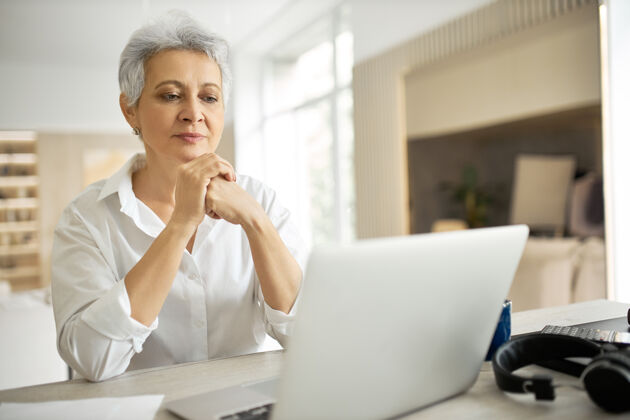 严肃侧视图：快乐的中年女商人 灰色短发 在时尚的办公室里用笔记本电脑工作 手放在键盘上 打字 分享好消息女性工作场所在线