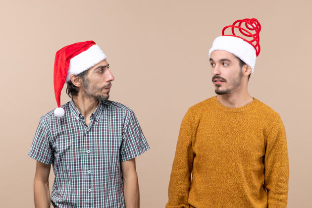 风景前视图两个戴着圣诞帽的家伙在米色孤立的背景下互相看着对方帽子两个伙计们男人