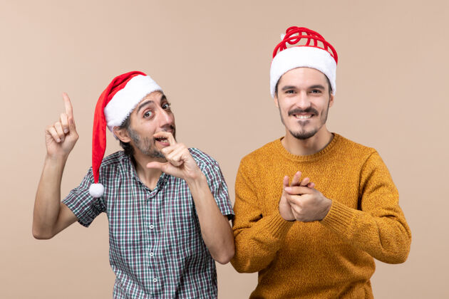 拿着正面图两个戴着圣诞帽的快乐的家伙 一个在展示什么 另一个在米色孤立的背景上双手合十米色两个帽子